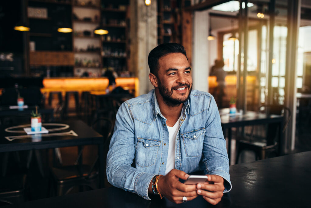 Young man in a cafe holding his phone and smiling.