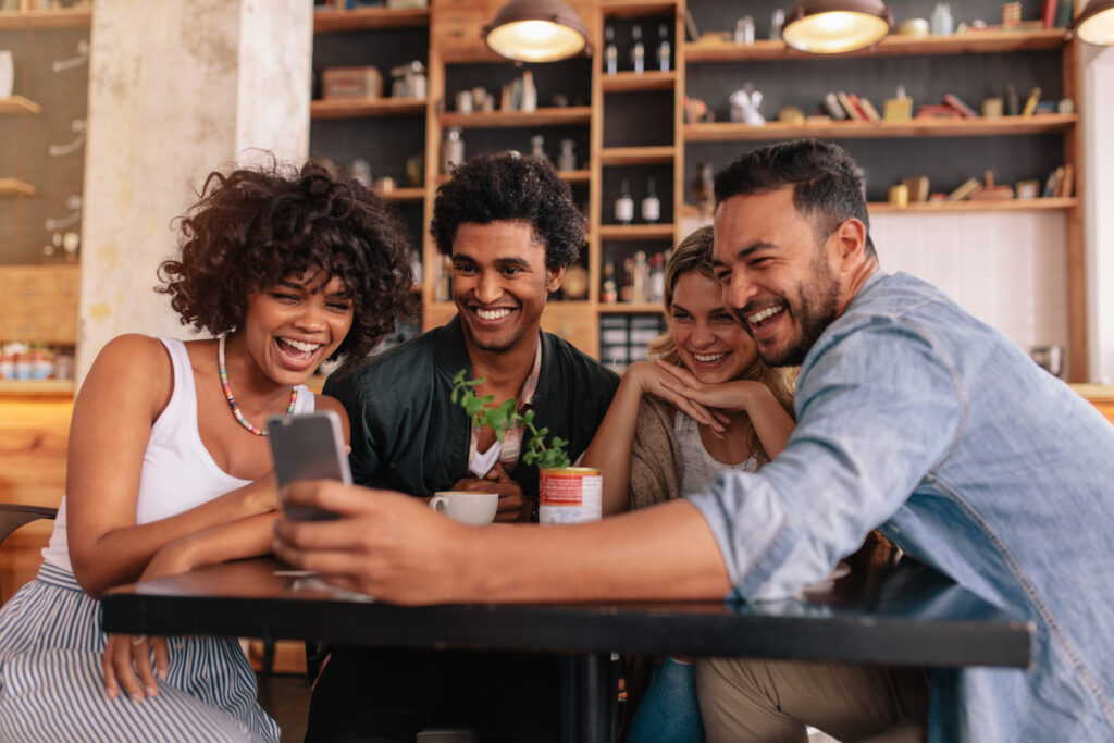 A group of friends sitting at a table in a cafe looking at a phone and laughing.
